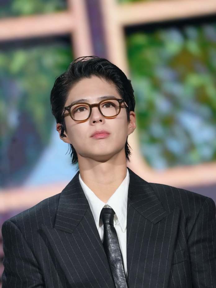 A young person wearing glasses and a pinstripe suit, looking thoughtfully upwards with a blurred background of greenery.