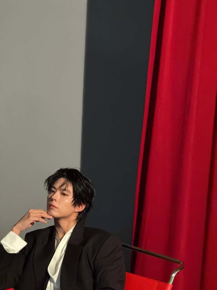 A contemplative person in a black suit sitting on a red chair with a dramatic red curtain in the background.
