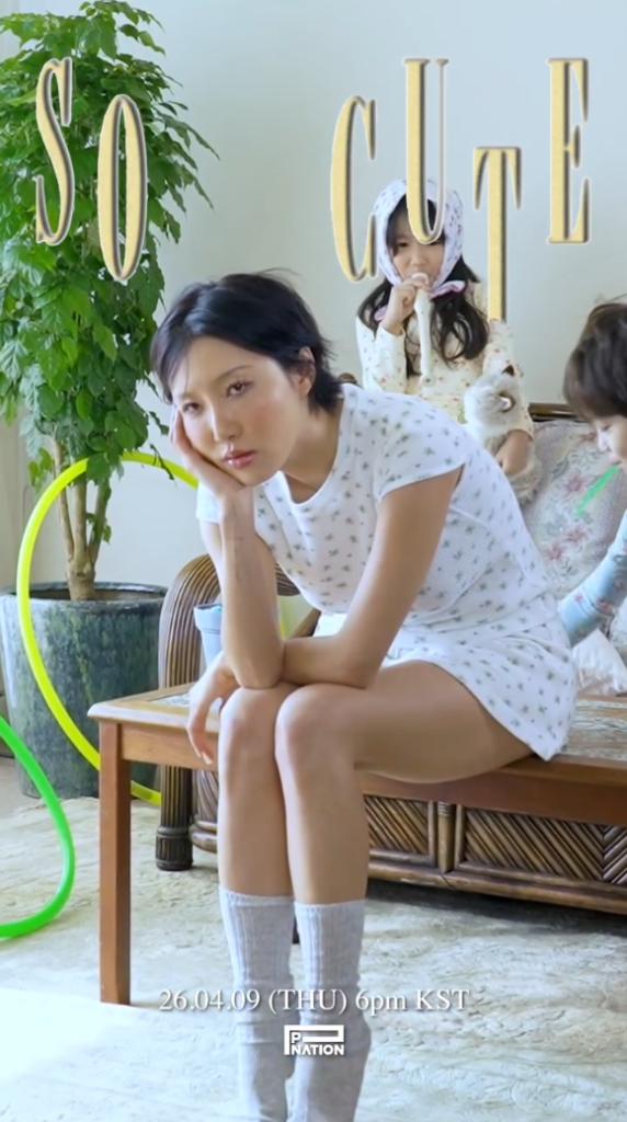 Hwasa sitting thoughtfully on a couch, wearing a patterned shirt and socks, with two children in the background, one holding a toy and looking at the camera, and the other observing, amidst indoor greenery.