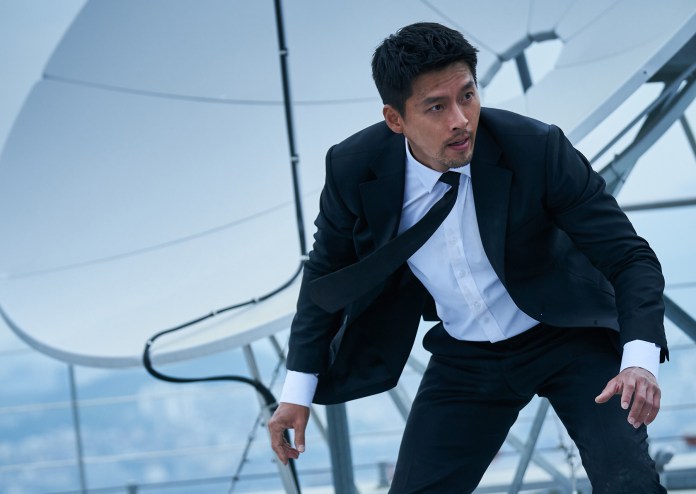Hyun Bin in a black suit and white shirt appears to be in a dynamic pose, possibly preparing to move or respond to a situation, against a backdrop of large white structures and a cloudy sky.