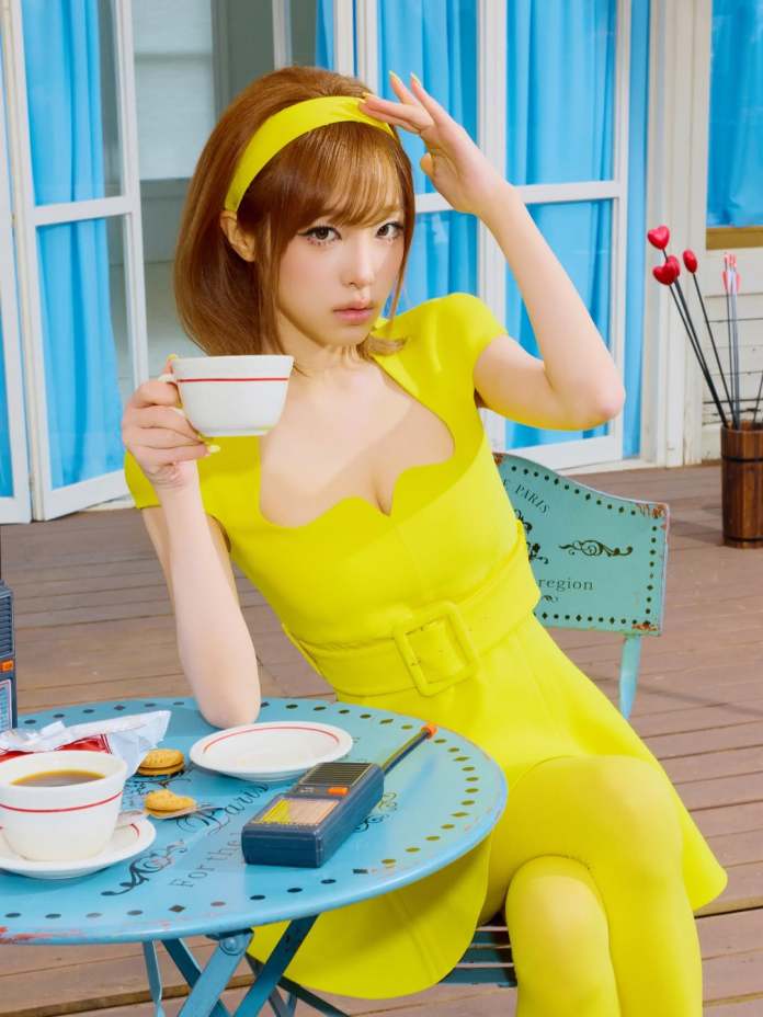 Choi Yena in a bright yellow dress and headband, sitting at a table with a cup of coffee, looking at the camera while gesturing with her hand.