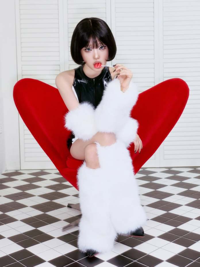 Choi Yena with a bob haircut sitting in a large red heart-shaped chair, wearing a black latex outfit and white fur accessories, playfully holding a red lollipop.