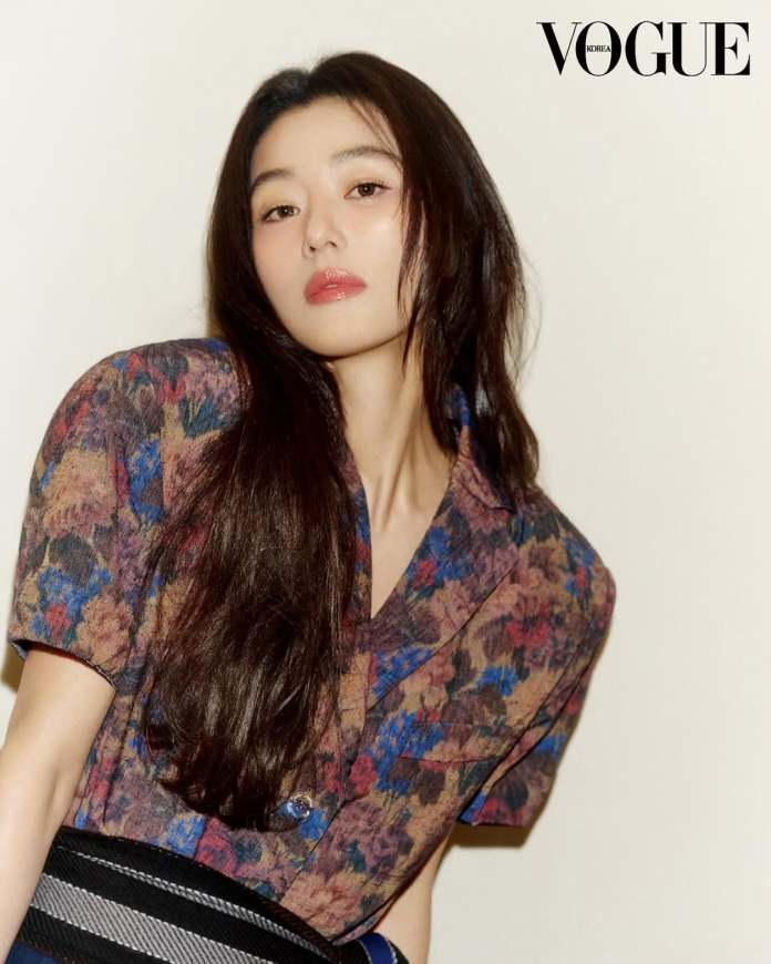 Jun Ji-hyun with long dark hair poses stylishly, wearing a patterned shirt and a wide belt, against a light background. The image is featured in Vogue Korea.
