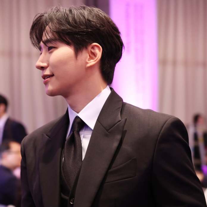 Lee Junho, looking away from the camera with a slight smile, set against a blurred background of a softly lit event.