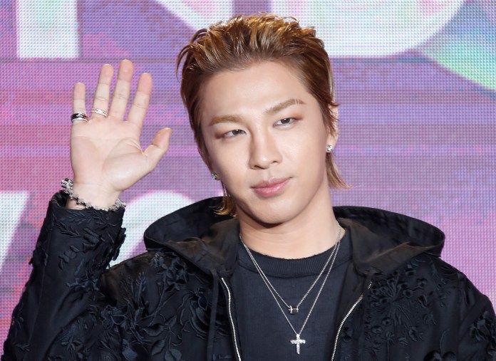 Taeyang with styled hair wearing a black jacket and multiple silver necklaces, raising one hand in a wave during a public appearance.