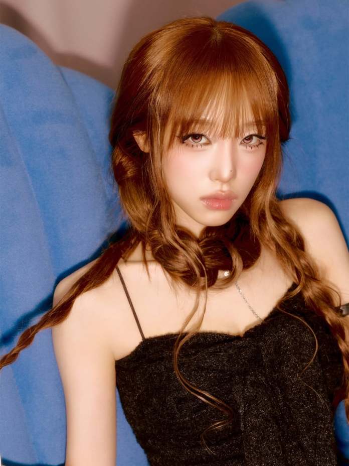 Choi Yena with long, wavy brown hair styled in two braids, wearing a black top, posing with a serious expression against a blue background.