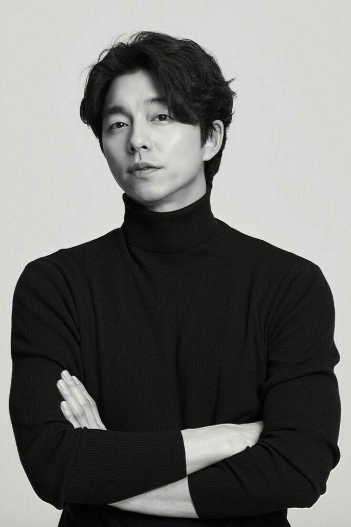 Gong Yoo with medium-length dark hair is posing confidently, wearing a black turtleneck sweater. He has his arms crossed and is looking directly at the camera against a plain white background.