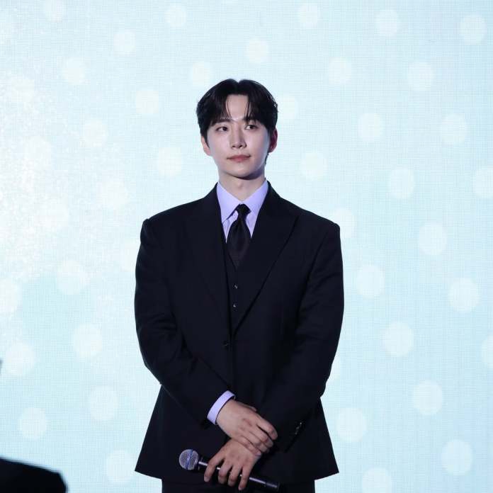 Lee Junho in a black suit stands on stage holding a microphone, with a soft blue background.