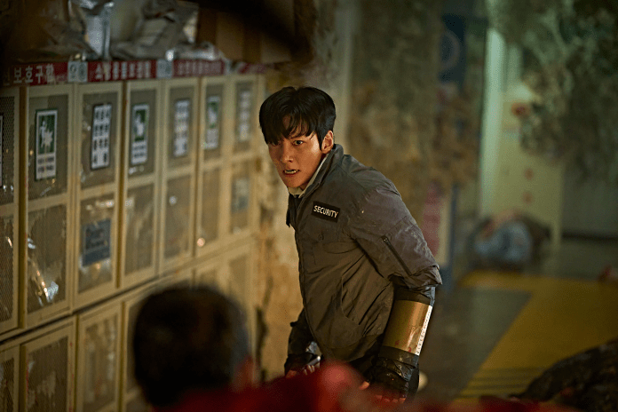 Ji Chang Wook, wearing a security uniform, looks intently towards an unseen figure while standing in a dimly lit hallway filled with lockers and disarray.