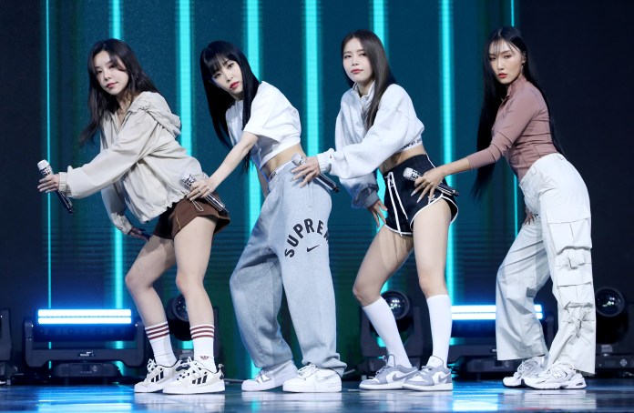 MAMAMOO members dancing in coordinated outfits on stage, holding microphones, with vibrant lighting in the background.