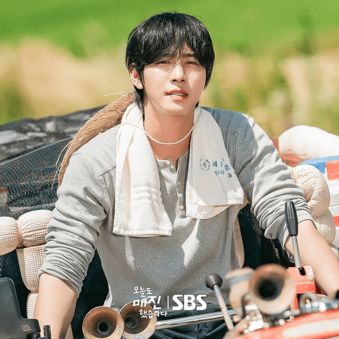 Ahn Hyo Seop with long black hair and a casual gray shirt sitting on a tractor, surrounded by agricultural scenery.