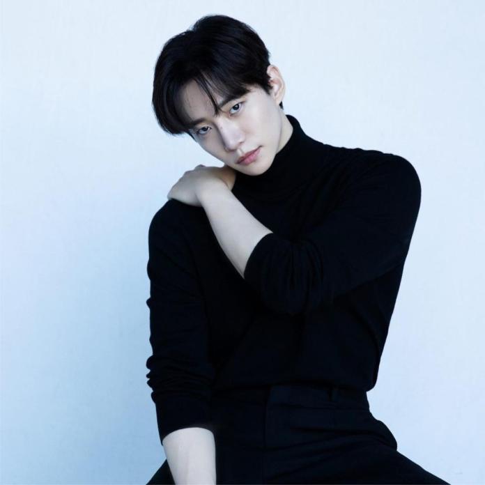 Lee Junho wearing a black turtleneck sits with one arm resting on his shoulder, gazing thoughtfully at the camera against a light blue background.