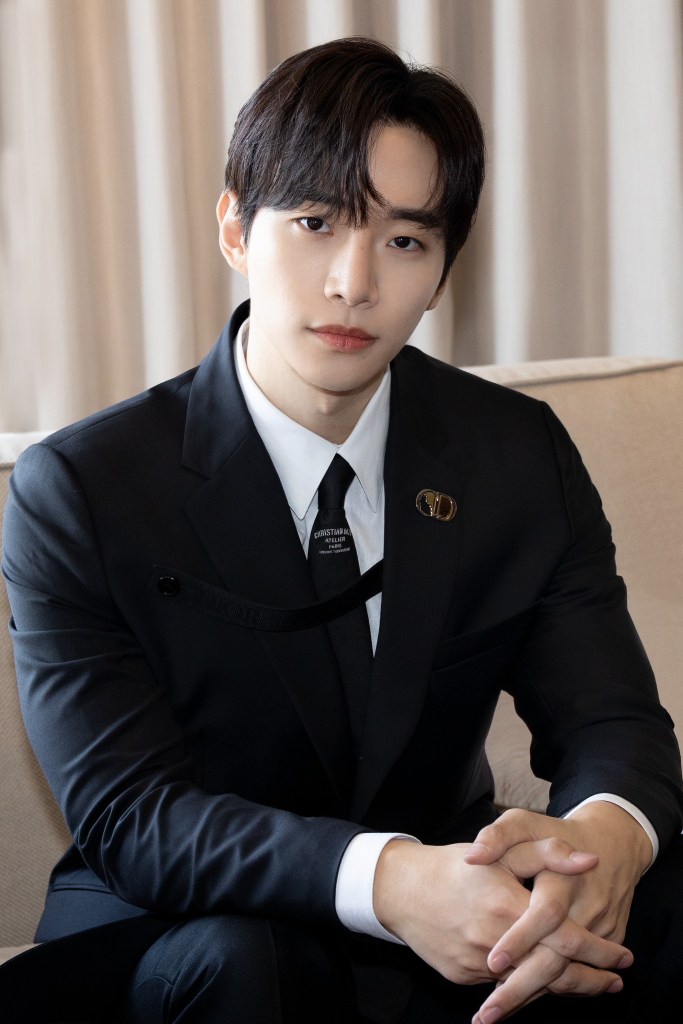Lee Junho in a tailored black suit with a white shirt and black tie, sitting on a beige sofa and looking directly at the camera with a serious expression.
