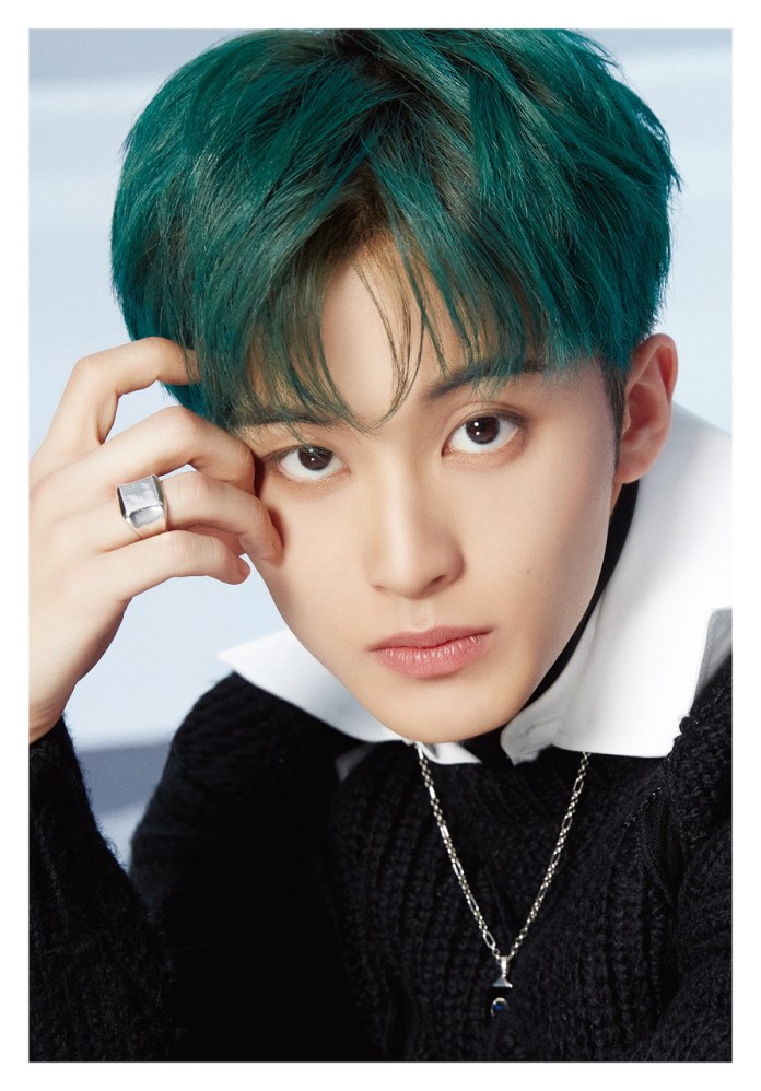 Mark Lee with teal hair, wearing a black sweater and a white collar. They are resting their hand on their face and staring directly at the camera.