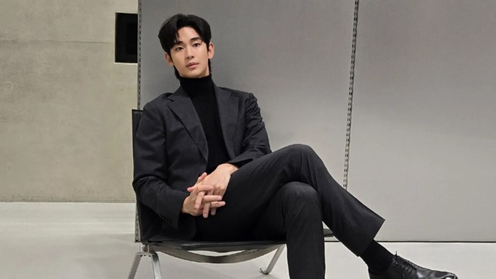 Kim Soo-hyun wearing a black turtleneck and a gray suit sits casually in a modern chair, with a minimalist backdrop.