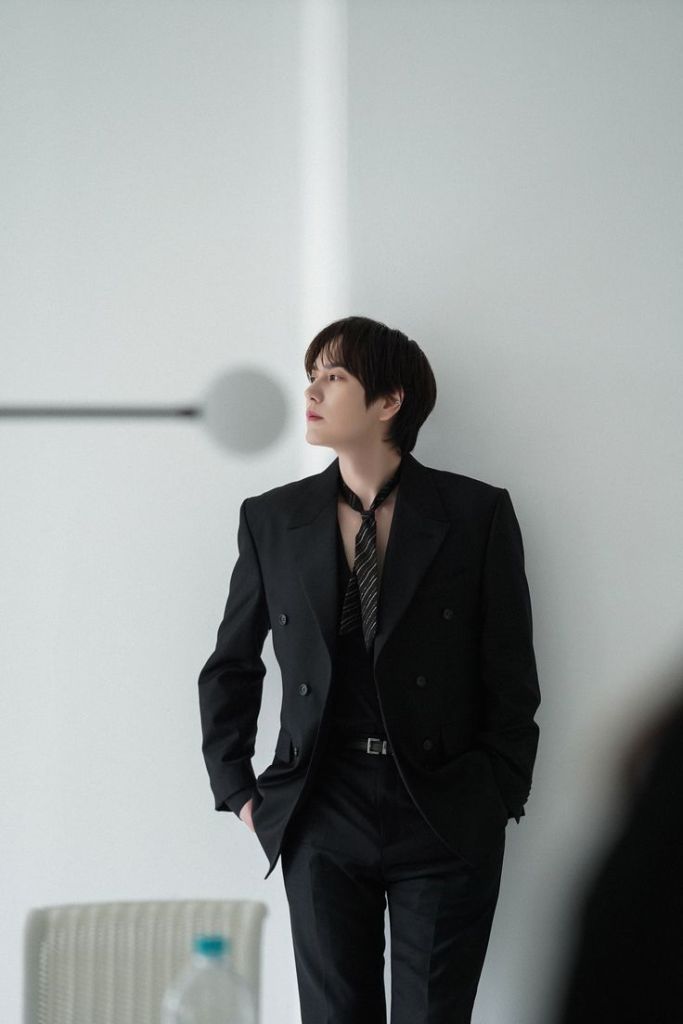 Kyuhyun dressed in a black suit with an open collar and a tie, leaning against a wall in a modern, minimalist setting.