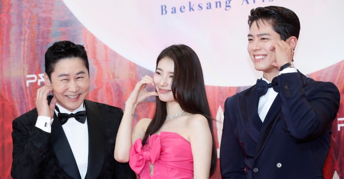Shin Dong-yup, Suzy, and Park Bo-gum posing at an event, with one man in a tuxedo, a woman in a pink dress, and another man in a suit, all smiling and making playful gestures.