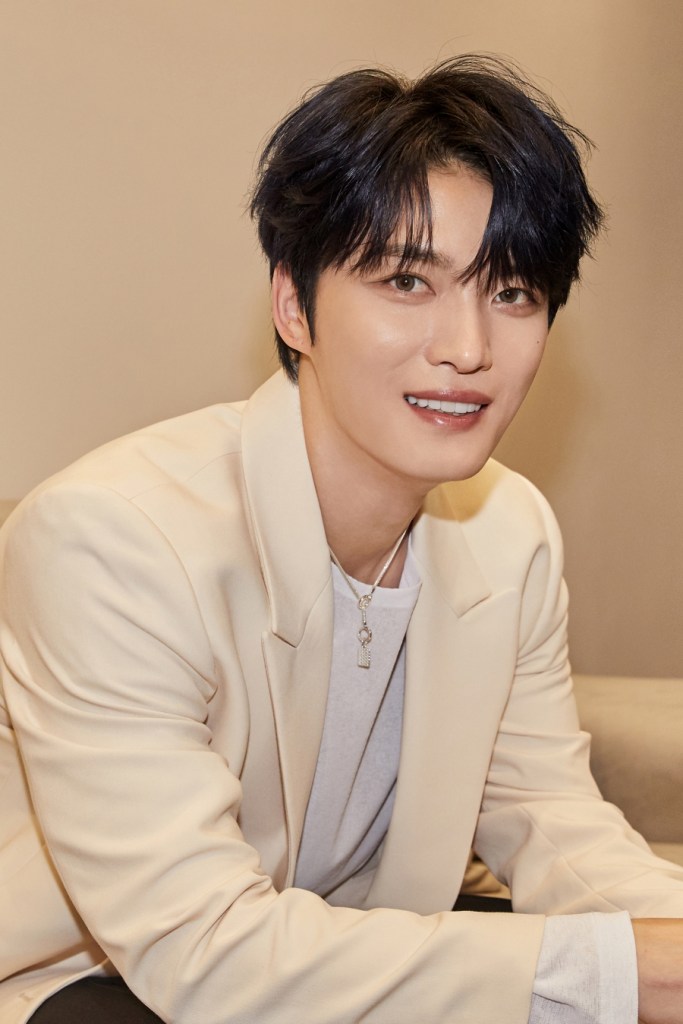 Kim Jae-joong with dark, tousled hair smiles at the camera while wearing a light-colored suit jacket over a white shirt. He appears relaxed, seated on a couch in a softly lit room.