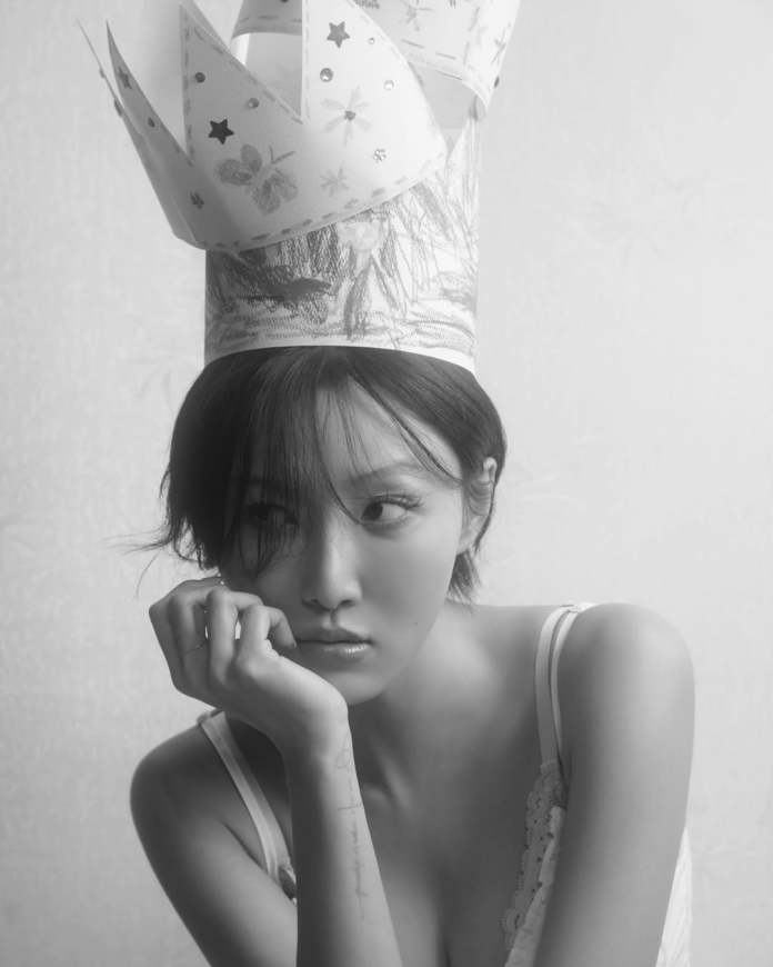 Hwasa with short hair resting her chin on her hand, wearing a decorative paper crown. The image is in black and white, featuring a soft background.