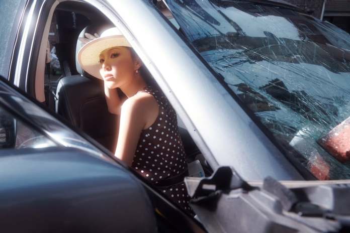 Hwasa wearing a polka dot dress and a wide-brimmed hat sits inside a car, gazing thoughtfully out the window. The scene includes a shattered windshield reflecting light.