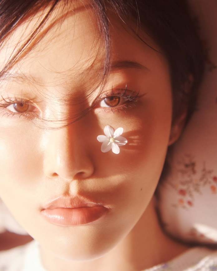 Close-up of Hwasa with brown eyes and natural makeup, featuring a small white flower placed on her cheek, illuminated by soft, warm lighting.