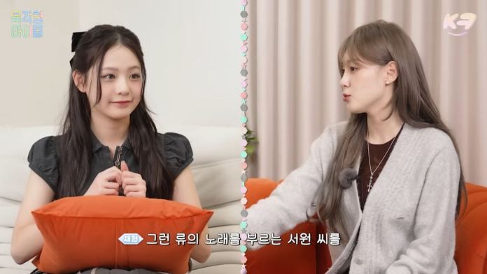 UNIS and Kep1er sitting on a sofa discussing, one holding an orange pillow and the other speaking animatedly, with a light-colored curtain in the background.