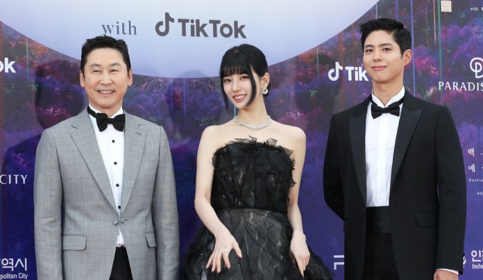 Shin Dong-yup, Suzy, and Park Bo-gum posed for a photo at a formal event, with a backdrop featuring TikTok branding. The first person on the left wears a gray tuxedo, the second person in the center wears a black dress, and the third person on the right is dressed in a black suit.