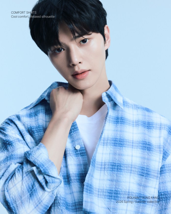 Song Kang posing confidently in a blue plaid shirt over a white t-shirt, with a soft blue background.