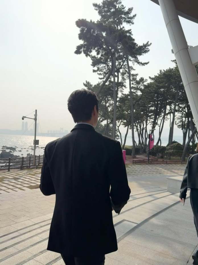 Lee Junho in a black coat walking away from the camera along a waterfront promenade surrounded by trees, with a hazy skyline in the background.