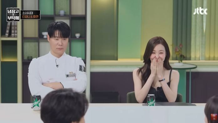 A man in a chef's uniform sits with arms crossed, looking pensive, while a woman in a black dress sits beside him, smiling and covering her mouth with her hands in surprise or laughter.