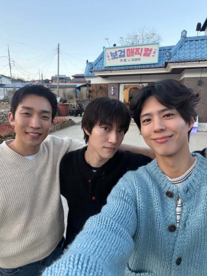 Park Bo-gum, Lee Sang-yi, and Kwak Dong-yeon pose for a selfie outside a building with a blue roof and a sign in Korean, wearing cozy sweaters in a rural setting.
