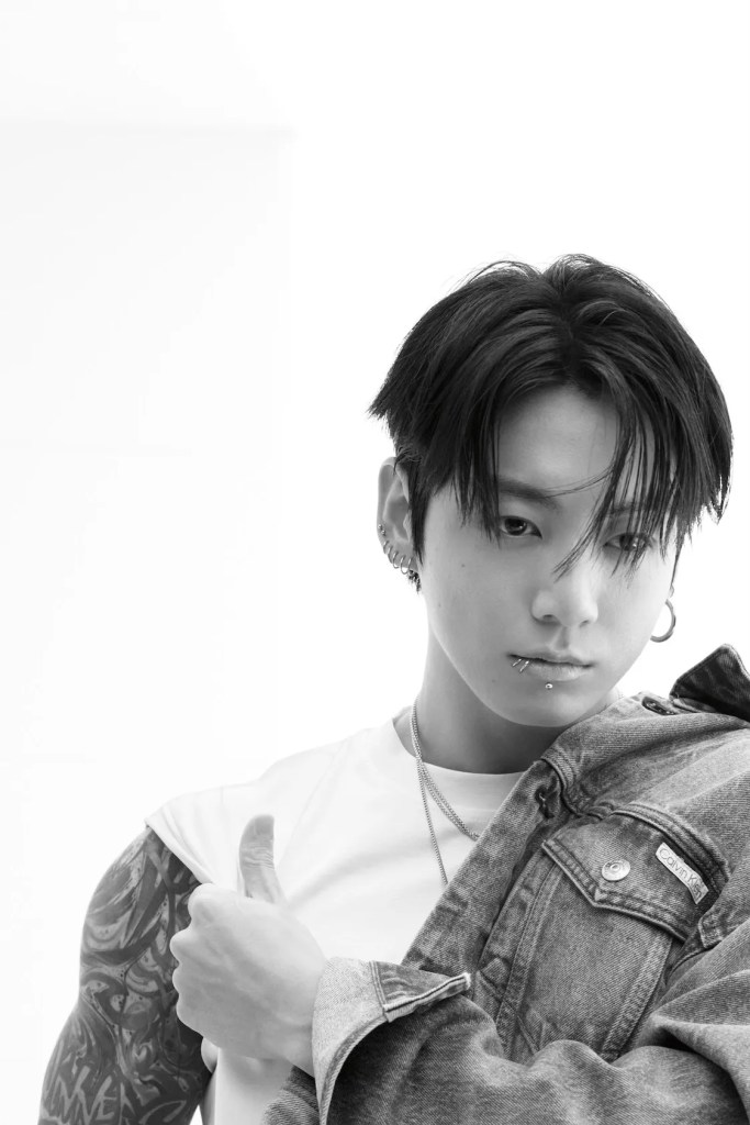 Jung kook with dark hair styled in a modern cut poses in a black and white image, wearing a white t-shirt and a denim jacket. He has multiple piercings and tattoos visible on his arm, and he is looking directly at the camera with a confident expression.