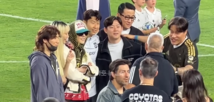 G-Dragon, Daesung and Son Heung-min posing together on a soccer field, smiling and socializing. In the background, a few children and other individuals are visible.