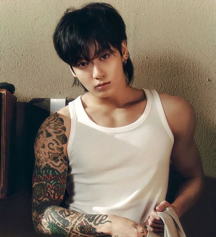 Jung Kook with long black hair and a sleeveless white shirt, showcasing intricate tattoos on their left arm, sitting against a textured wall.