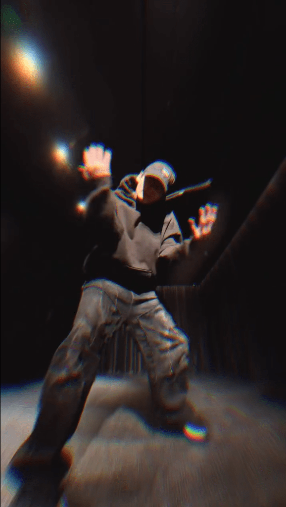 Jungkook wearing a black hoodie and cap is striking a dynamic pose in a dark room, with dramatic lighting effects highlighting their hands.