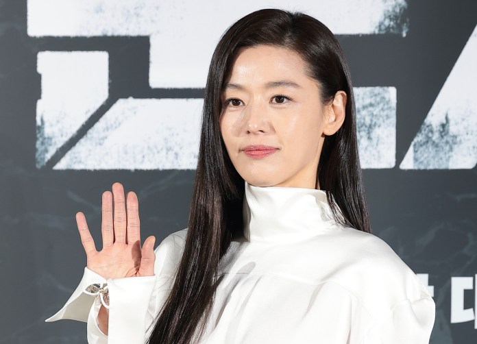 Jun Ji-hyun with long black hair wearing a white high-collared outfit, waving her hand at an event backdrop.