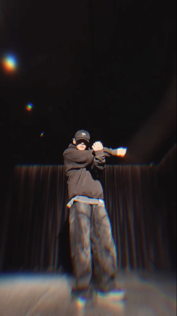 Jungkook performing on stage, wearing a black hoodie and cap, with a dramatic pose against a dark background.