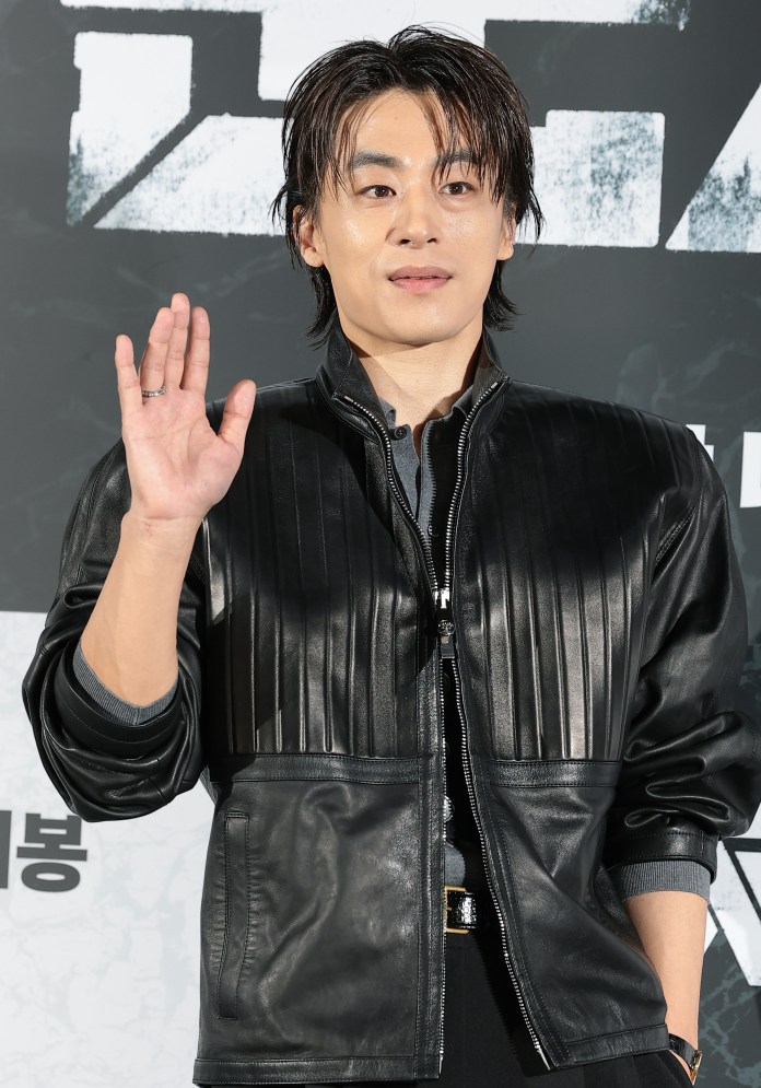 Koo Kyo-hwan wearing a black leather jacket and greeting with a hand wave at a press event.