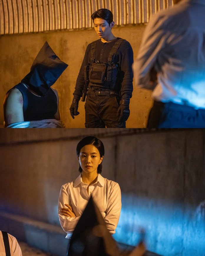 A tense scene featuring Dex in black tactical clothing standing over another man who is bound and wearing a black hood Lee Seol in a white shirt stands off to the side, arms crossed, observing the situation.
