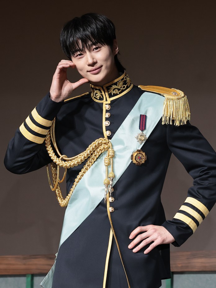  Byeon Woo-seok  in a historical military-style uniform with gold embellishments and a light blue sash, posing with a friendly smile and hand on his chin.