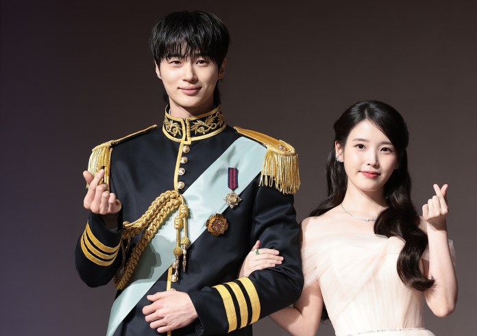  Byeon Woo-seok in a military-style uniform with gold accents and a sash poses next to IU in a light-colored evening gown, both making small heart gestures with their hands against a neutral backdrop.