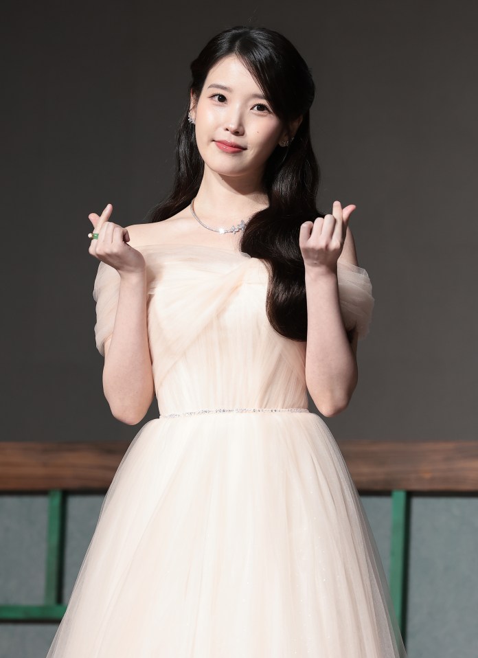 IU wearing a strapless, pale pink gown with a tulle skirt, smiling and making a heart gesture with her hands.