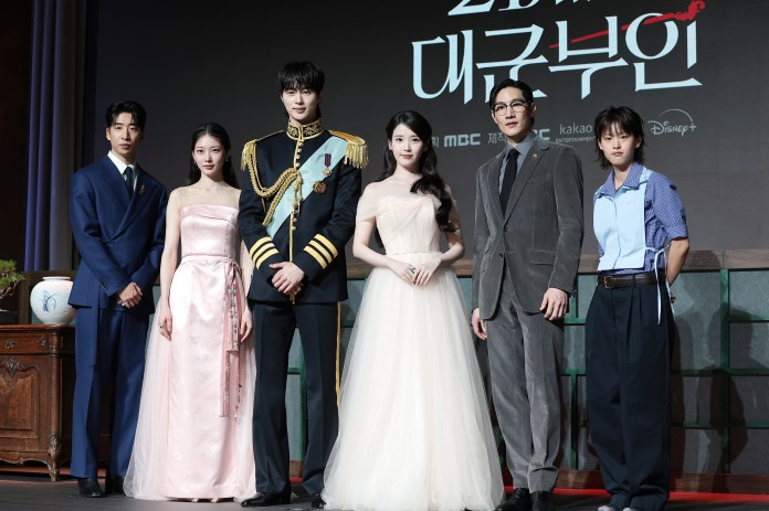 IU, Byeon Woo-seok, Noh Sang-hyun, Gong Seung-yeon, Lee Yeon posing for a photo at a promotional event. They are dressed in formal attire, with one person in a military outfit. The backdrop displays the title of a show in Korean.