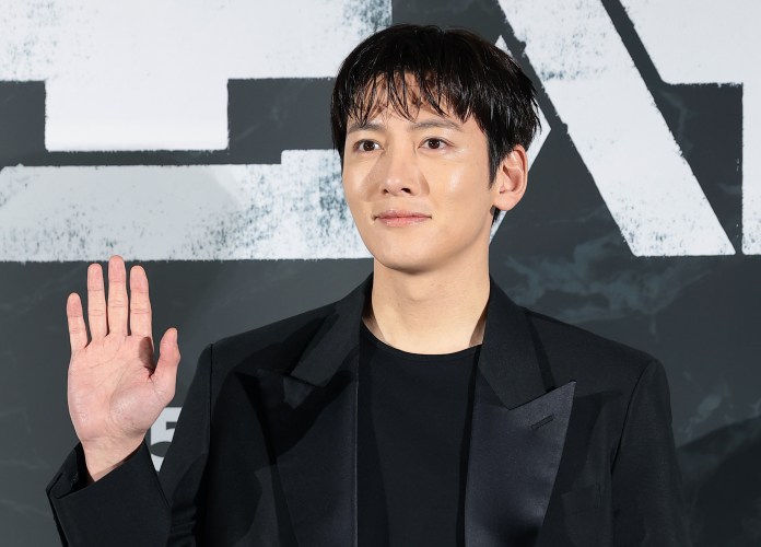  Ji Chang-wook in a black blazer waving at an event, standing in front of a gray backdrop with abstract designs.