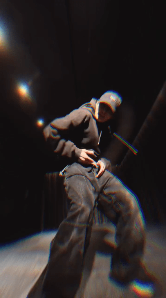 Jungkook in a dark space wearing a hoodie and cap, performing a pose with hands on their waist.