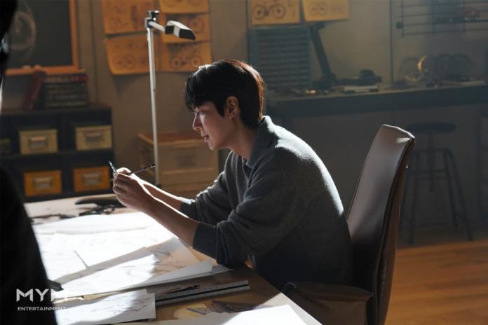 Lee Min-ho sitting at a desk, focused on drawing or sketching, surrounded by a well-lit and creative workspace filled with design tools and papers.
