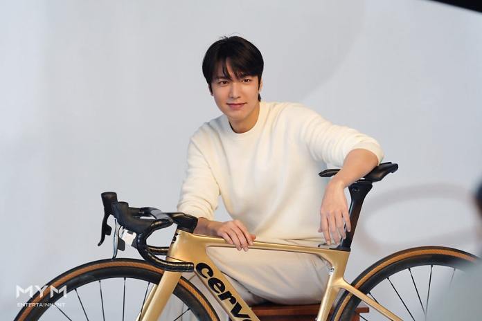 Lee Min-ho dressed in a white sweater and pants, sitting on a stool beside a gold road bicycle, smiling at the camera.