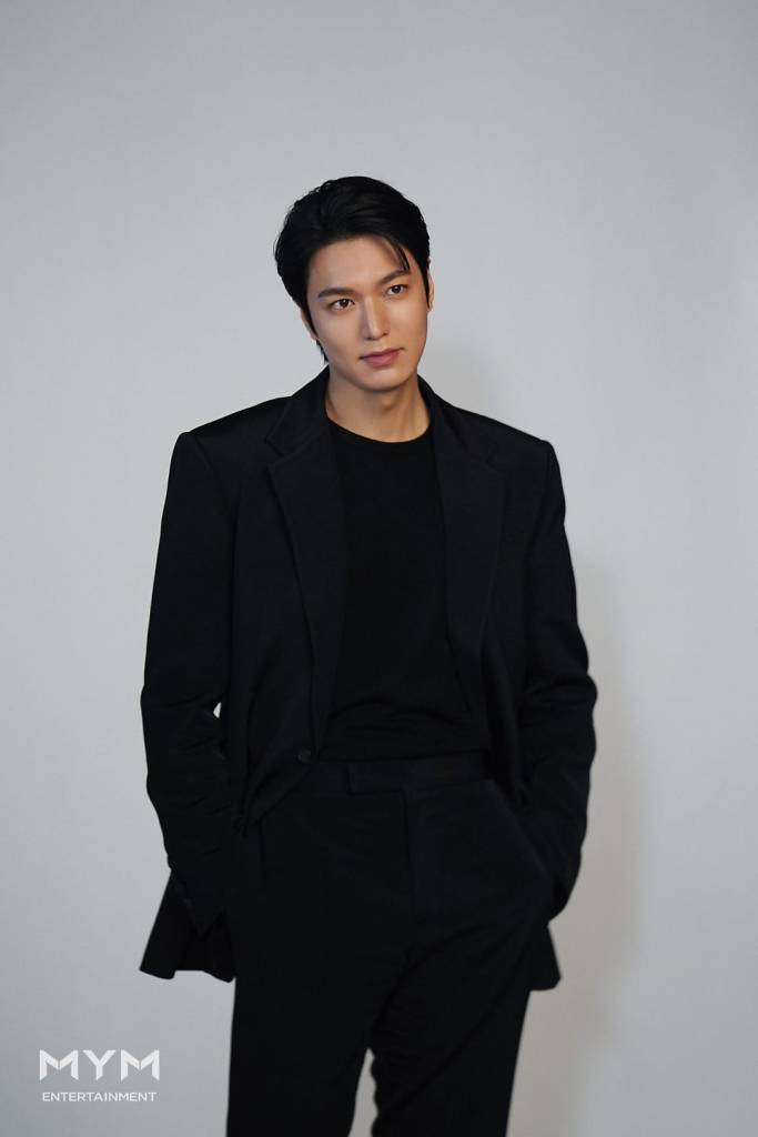 Lee Min-ho in a black suit stands confidently against a neutral background, looking slightly to the side with a subtle smile.