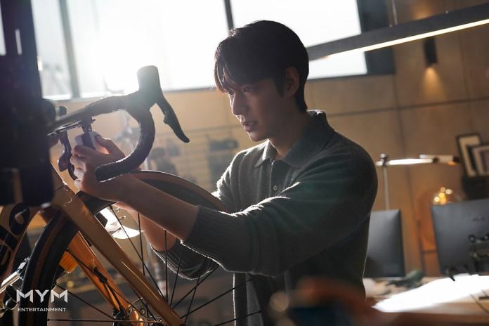 Lee Min-ho inspecting and adjusting a bicycle in a well-lit workspace, with desktop computers and office items in the background.