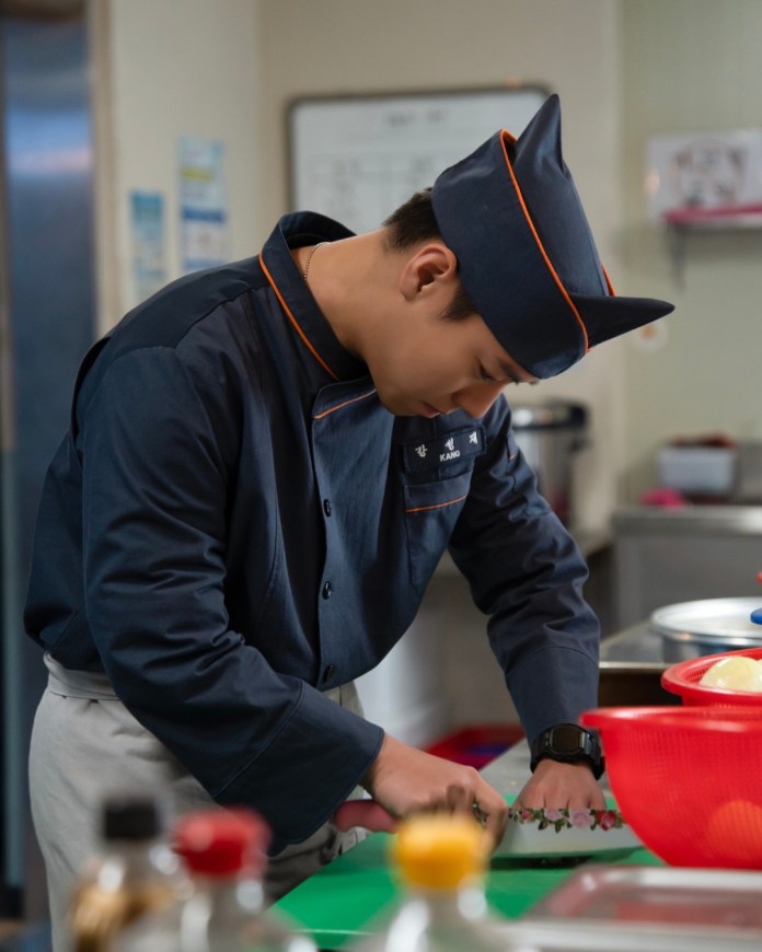 Park Ji-hoon wearing a dark blue uniform and hat is focused on chopping vegetables on a green cutting board in a kitchen setting.